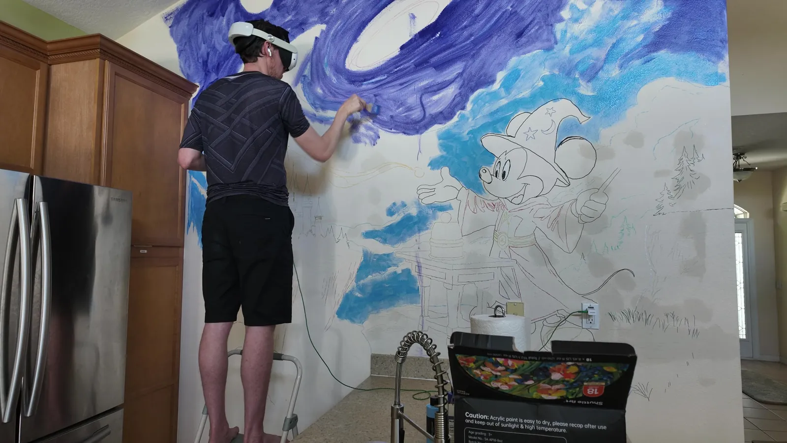 Stefan on a step stool painting the purple sky of a mural, with the rest of the design still in outline