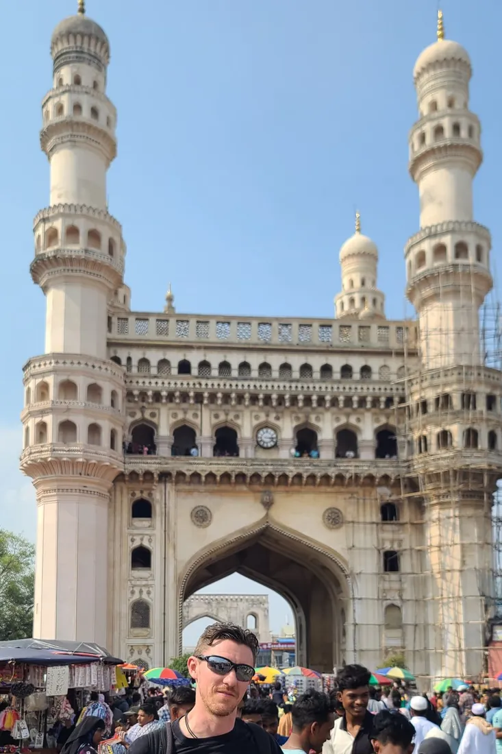 Stefan in sunglasses in front of the Charminar, a white monument with four minarets, on a busy market street